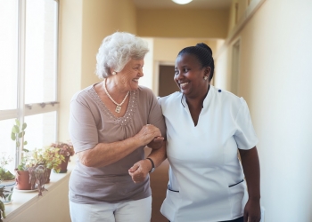 Portrait of smiling home caregiver and senior woman walking together through a corridor. Healthcare worker taking care of elderly woman.